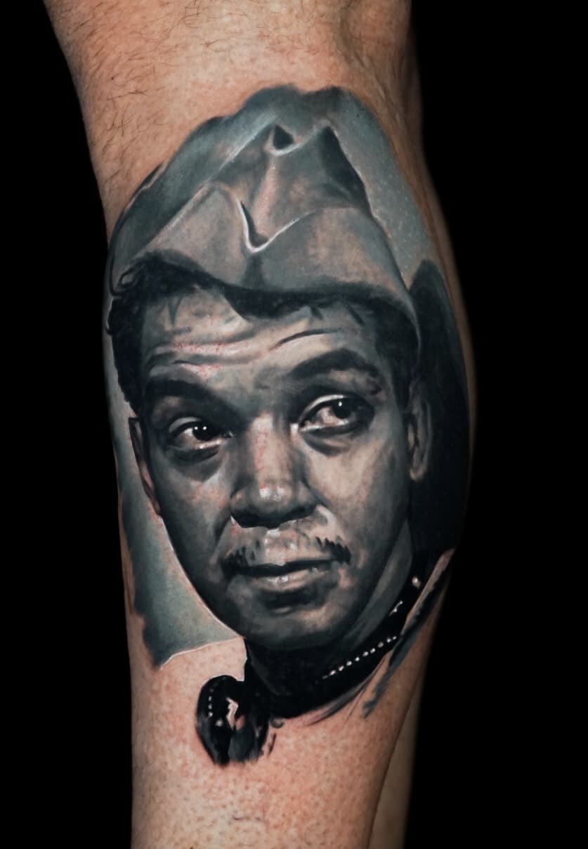 Jose Carlos 18 A grayscale tattoo of a man with a mustache and a hat, wearing a beaded necklace, on a person's arm.