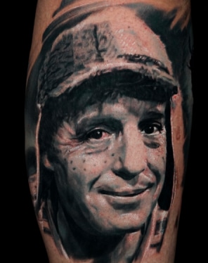 Home 11 A black and white tattoo of a smiling man with freckles wearing a worn cap with ear flaps.
