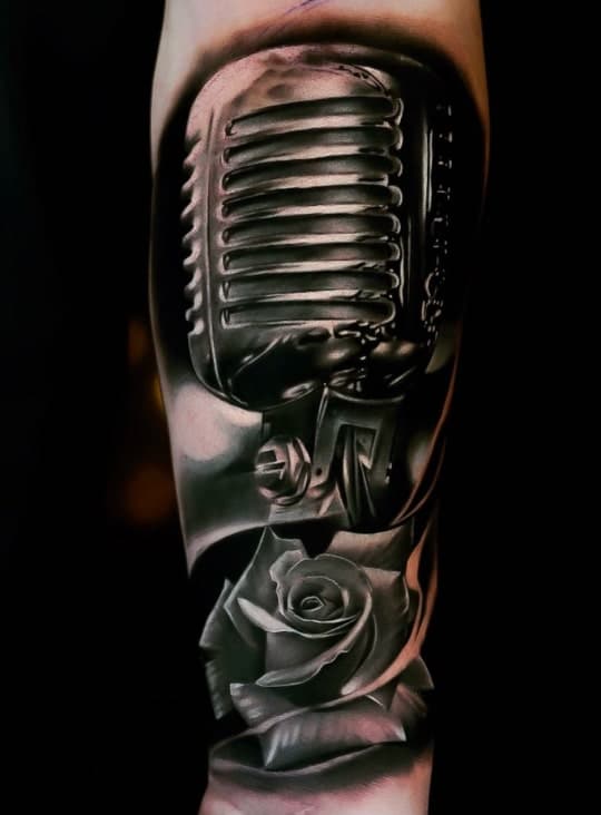 A black and white tattoo of a vintage microphone with a rose and leaves below it, on a forearm.