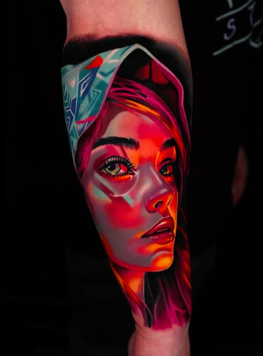 Home 9 A vibrant tattoo of a woman's face with pink hair, green eyes, and a blue patterned head covering.