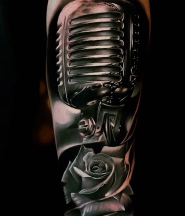 A black and white tattoo of a vintage microphone with a rose at its base on a person's arm.