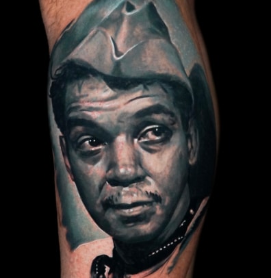 A black and white portrait tattoo of a man with a mustache and a cowboy hat on a person's arm.