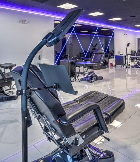 A modern tattoo parlor with white walls and dark ceiling featuring a black reclining chair and purple neon lights.