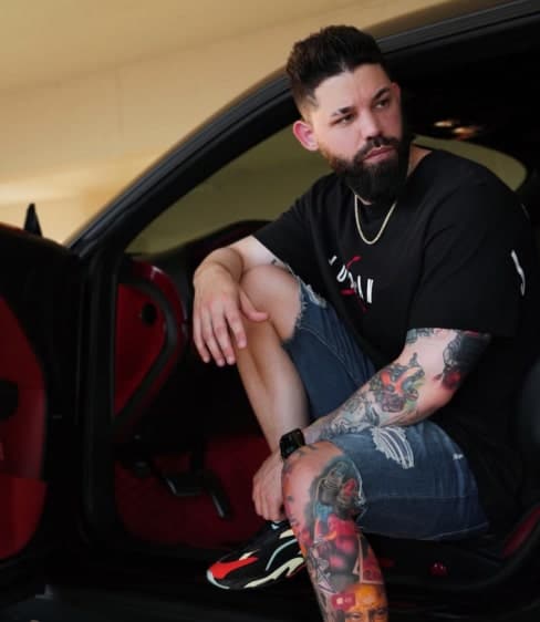 A bearded man with tattoos on his arms and legs sits in a car with a black and red interior.