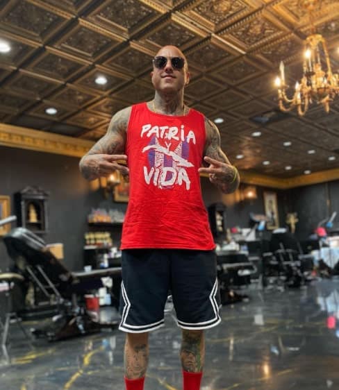 A heavily tattooed man in sunglasses, a red "PATRIA Y VIDA" tank top, and black shorts poses in a tattoo parlor.