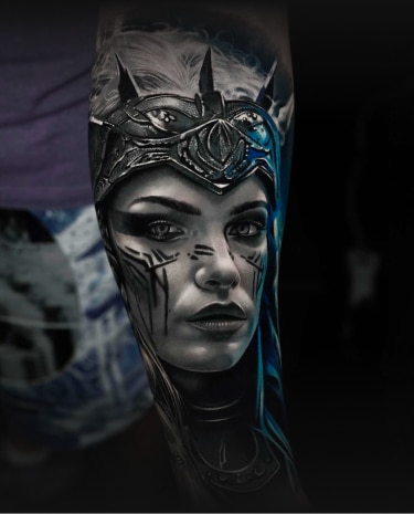 A detailed tattoo of a woman with a spiked crown, tribal face paint, and blue hair on an arm.