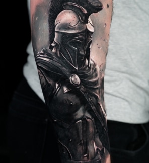Home 13 A detailed black and gray tattoo of a Spartan warrior in armor and helmet on a person's forearm.