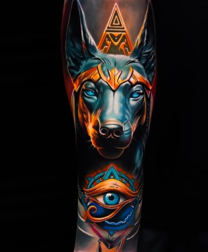Home 17 A vibrant tattoo on an arm depicts Anubis with glowing blue eyes and a gold headdress, above the Eye of Horus.