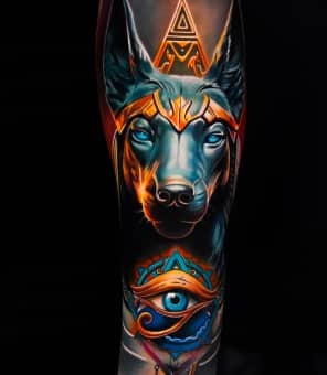 A colorful tattoo of a blue-eyed Anubis with a golden headdress and a blue Eye of Horus below.