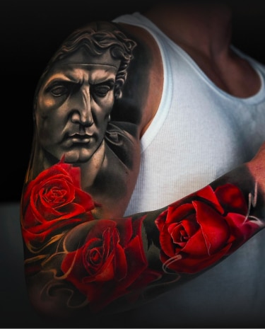 A man's arm is tattooed with a grayscale classical bust and several vibrant red roses.