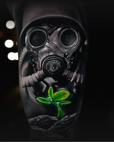 A black and white tattoo of a gas mask and helmet with a glowing green plant growing below it.