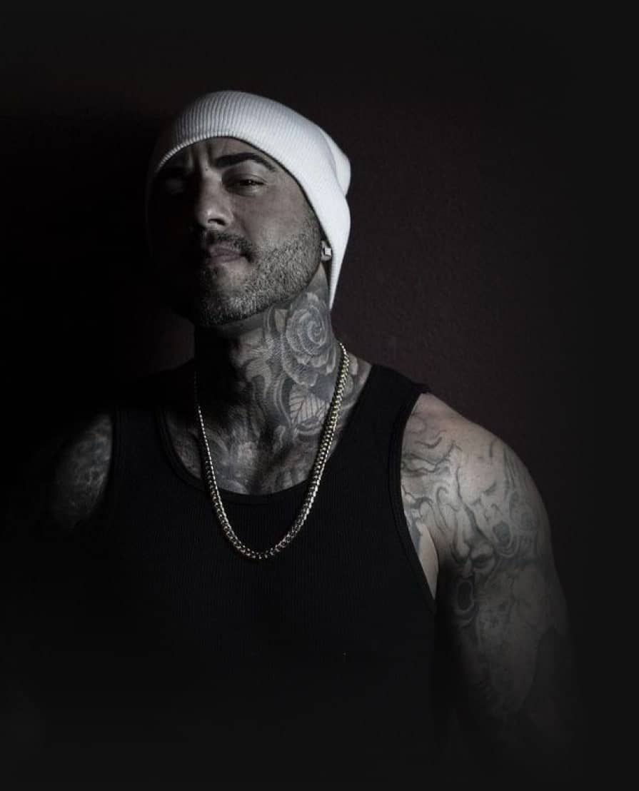 A man in a white beanie and black tank top with a gold chain and neck tattoos looks forward.