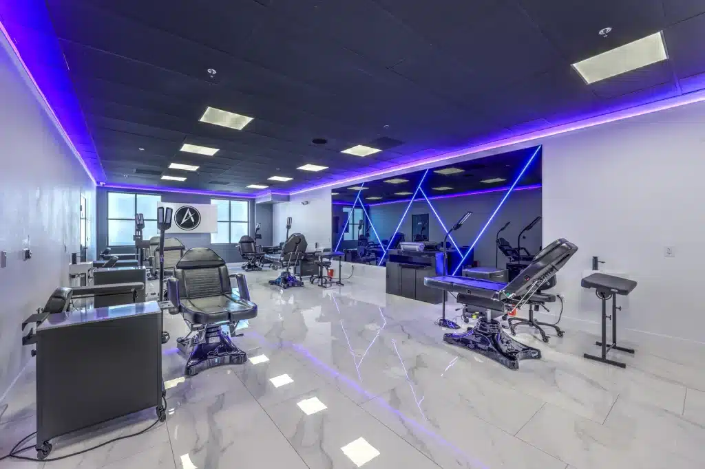 A modern tattoo studio with multiple black leather chairs, white marble floors, and purple LED strip lighting.