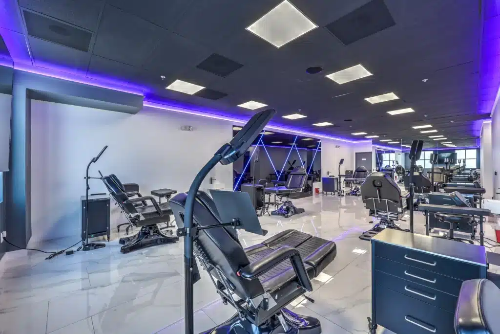 A spacious tattoo parlor features multiple black recliners, bright white tile floors, and purple LED lighting.