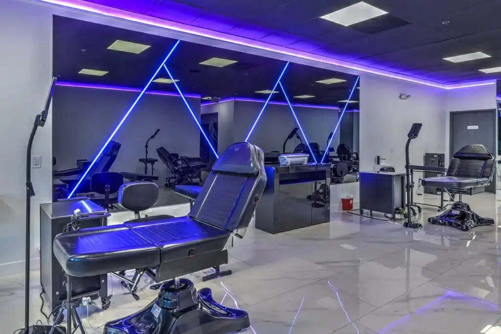 A modern tattoo parlor with black chairs, white marble floors, and mirrors reflecting blue neon lights.