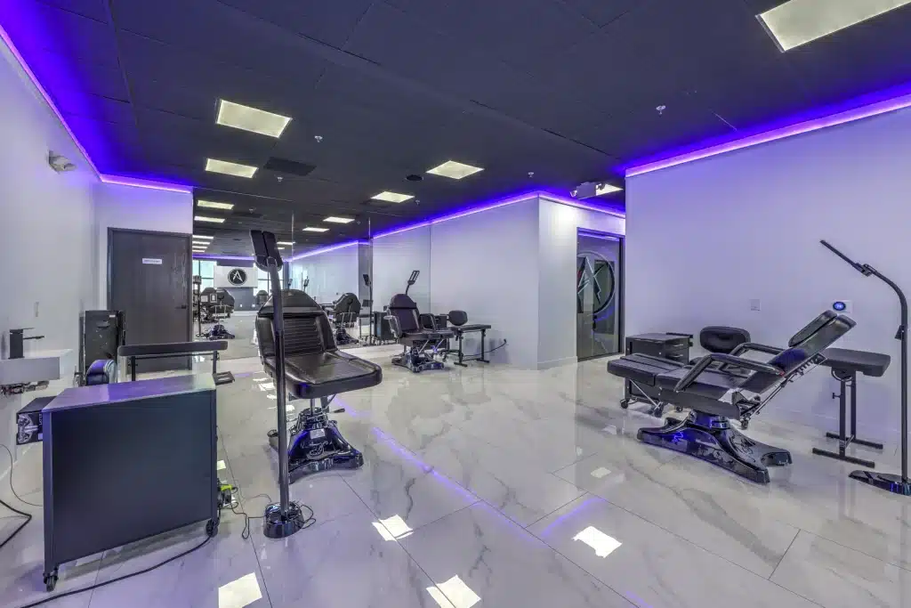 A modern tattoo studio with black chairs, white marble floors, and purple LED lighting along the ceiling.