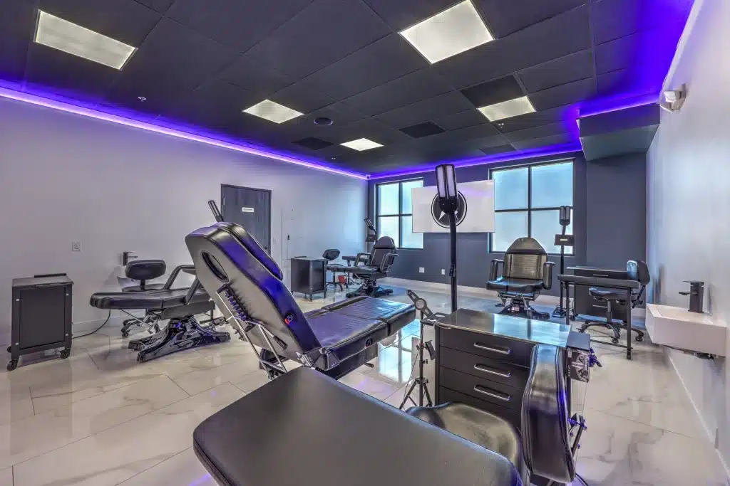 A modern tattoo studio with multiple black adjustable chairs and tables, illuminated by purple LED strips.