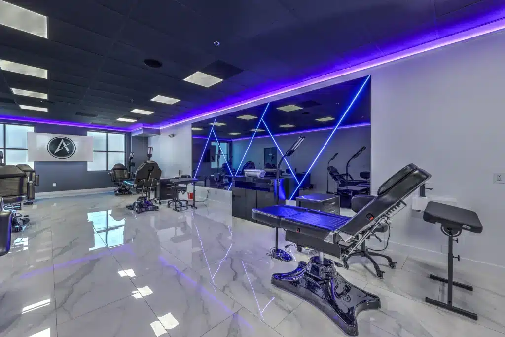 A modern tattoo parlor features black reclining chairs, white marble floors, and purple and blue neon lights.