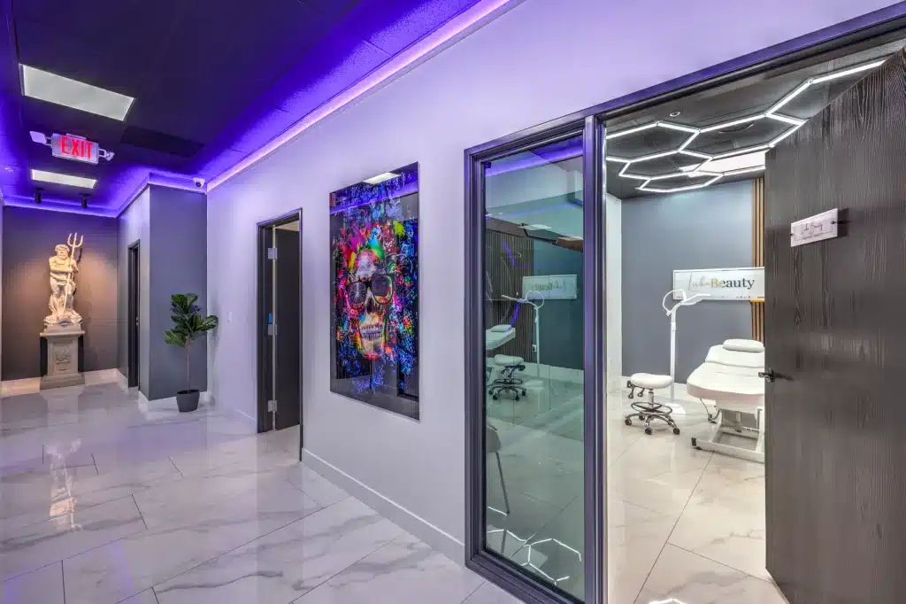 A hallway with purple lighting leads to a treatment room visible through a glass door, featuring a "Lash Beauty" sign.