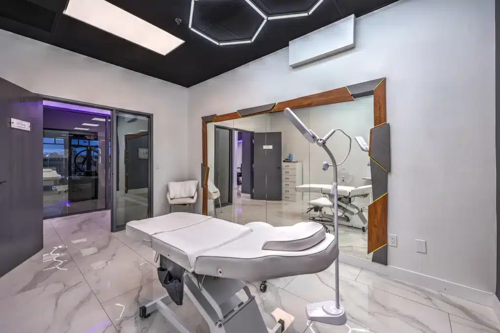 A modern treatment room with a black ceiling, white walls, a large mirror, and a white examination bed.