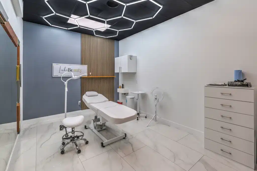 A modern beauty treatment room with a cream bed, white rolling stool, and "Lulu Beauty" sign on a gray wall.