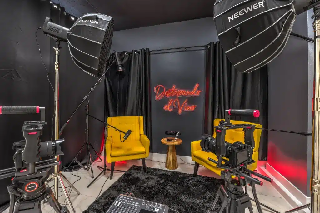 A dark studio with two yellow chairs, cameras, microphones, and "Destapando el Vino" in red neon on the wall.