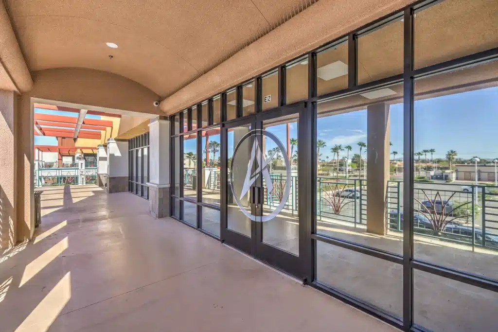 A long, covered outdoor walkway with a beige ceiling and floor, leading to glass doors with a white logo.