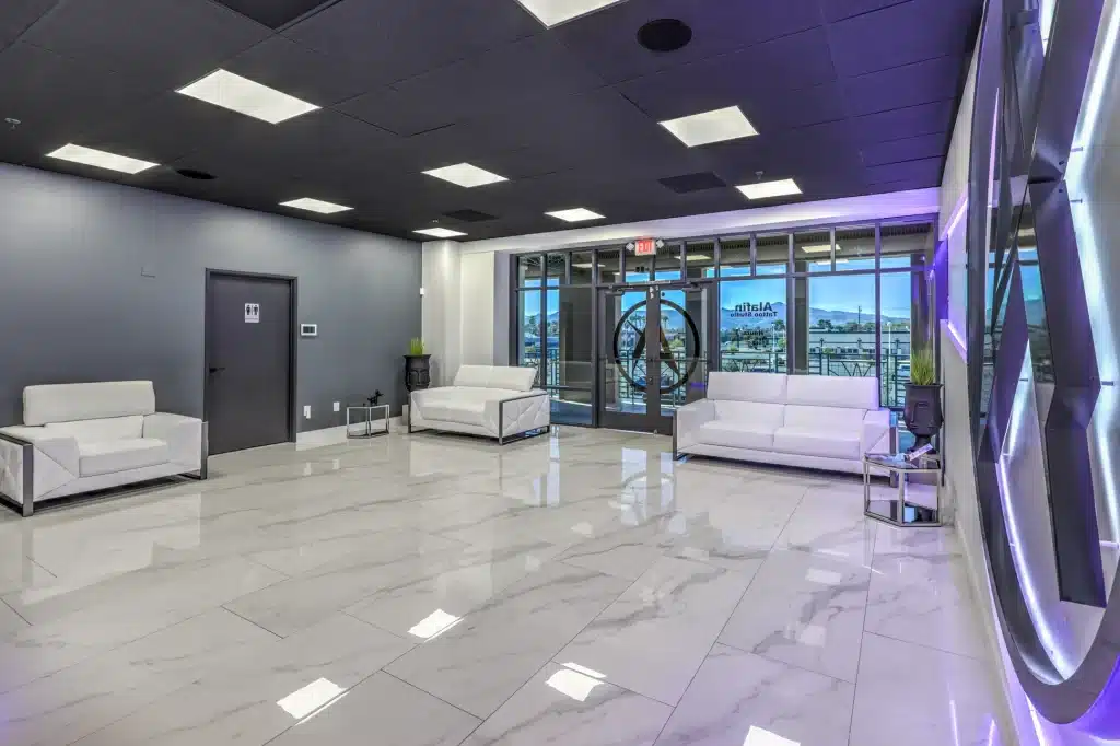 A modern lobby with white leather sofas, marble floors, a dark ceiling with square lights, and a glass door.