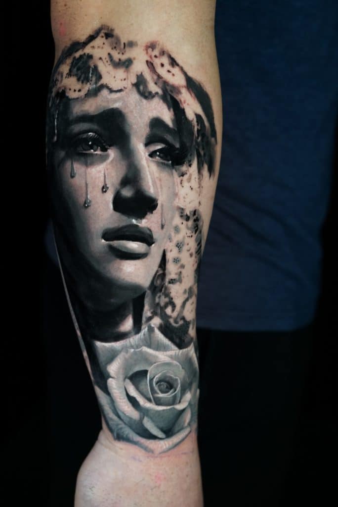 A black and white tattoo on an arm depicts a crying woman's face with a rose below it.