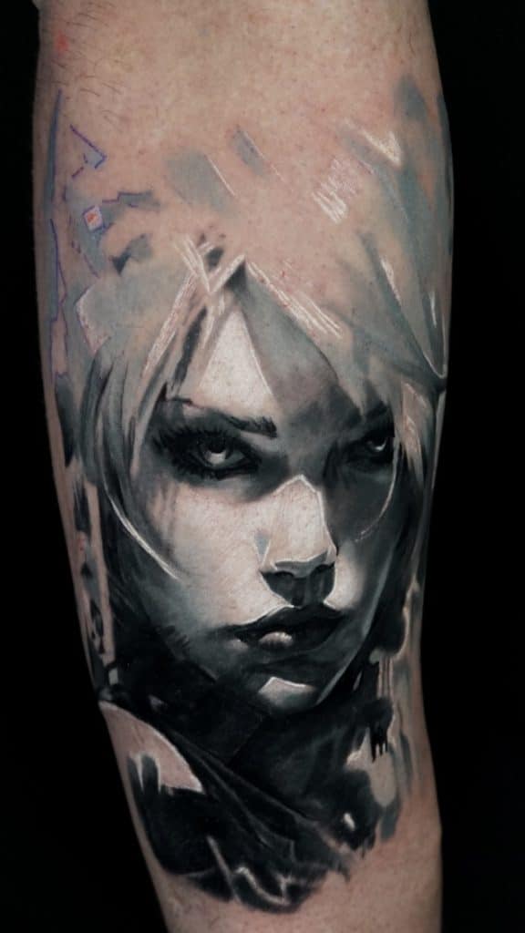 A grayscale tattoo of a woman's face with light hair, dark eyes, and a serious expression on an arm.