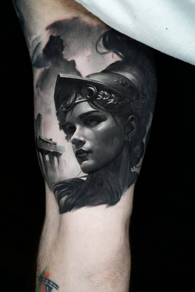 A black and white tattoo on an arm depicts a woman in a helmet with ancient ruins in the background.
