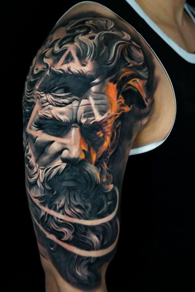 A detailed tattoo on an arm depicts a stone-like face with a beard, partially obscured by orange flames.