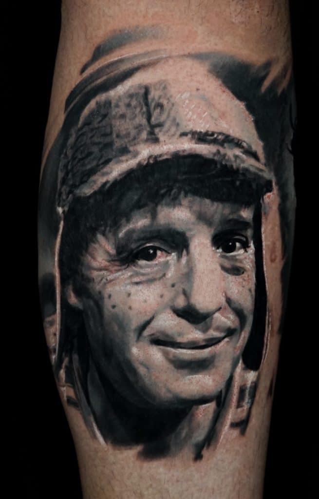A black and white realism tattoo of a smiling man with a cap, freckles, and wrinkles on a person's leg.