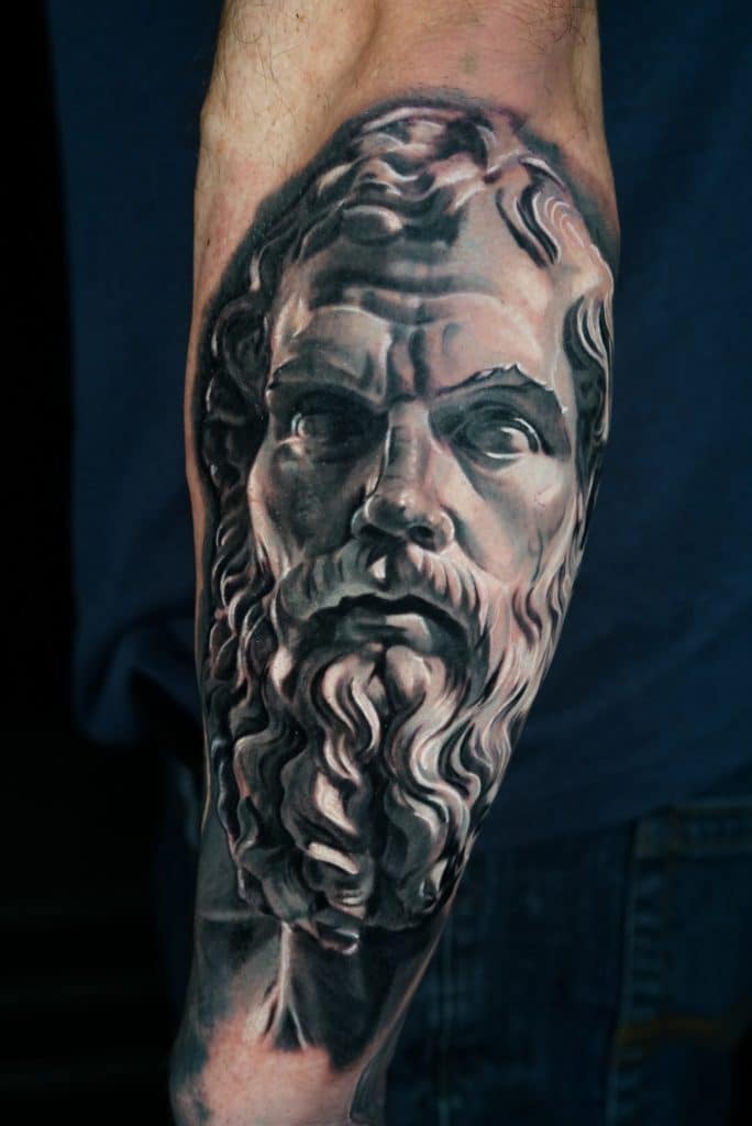 A grayscale tattoo on an arm depicts a classical bust of a man with a beard and wavy hair.