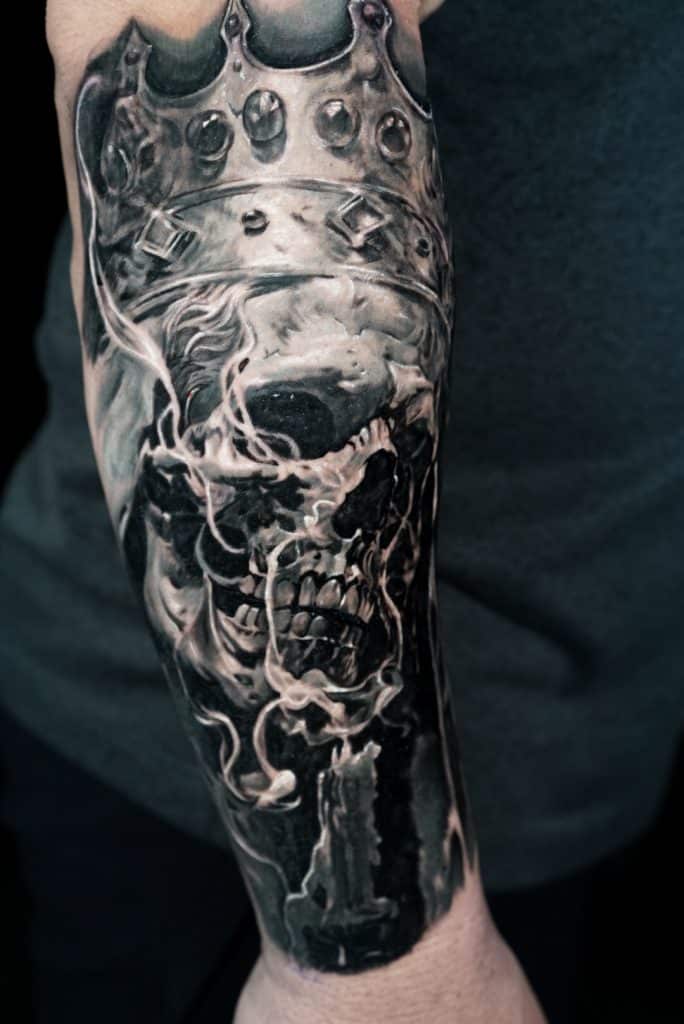 A black and white tattoo on a person's forearm depicts a skull wearing a crown, with smoke and a candle.