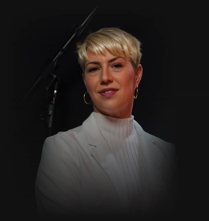 A woman with short blonde hair, gold hoop earrings, and a white turtleneck smiles against a dark background.