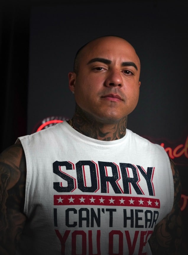 A bald, tattooed man with a gold chain wears a white sleeveless shirt that says "SORRY I CAN'T HEAR YOU OVER".