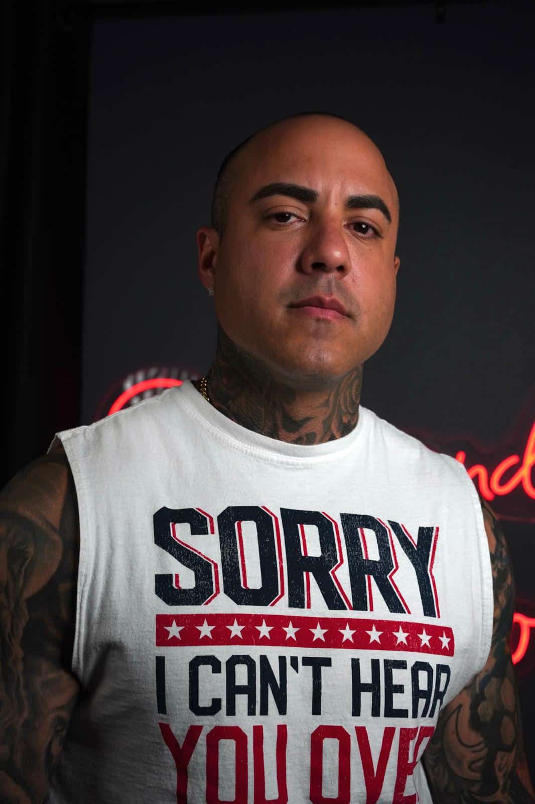 A bald man with neck and arm tattoos wears a white tank top that says "SORRY I CAN'T HEAR YOU OVER."