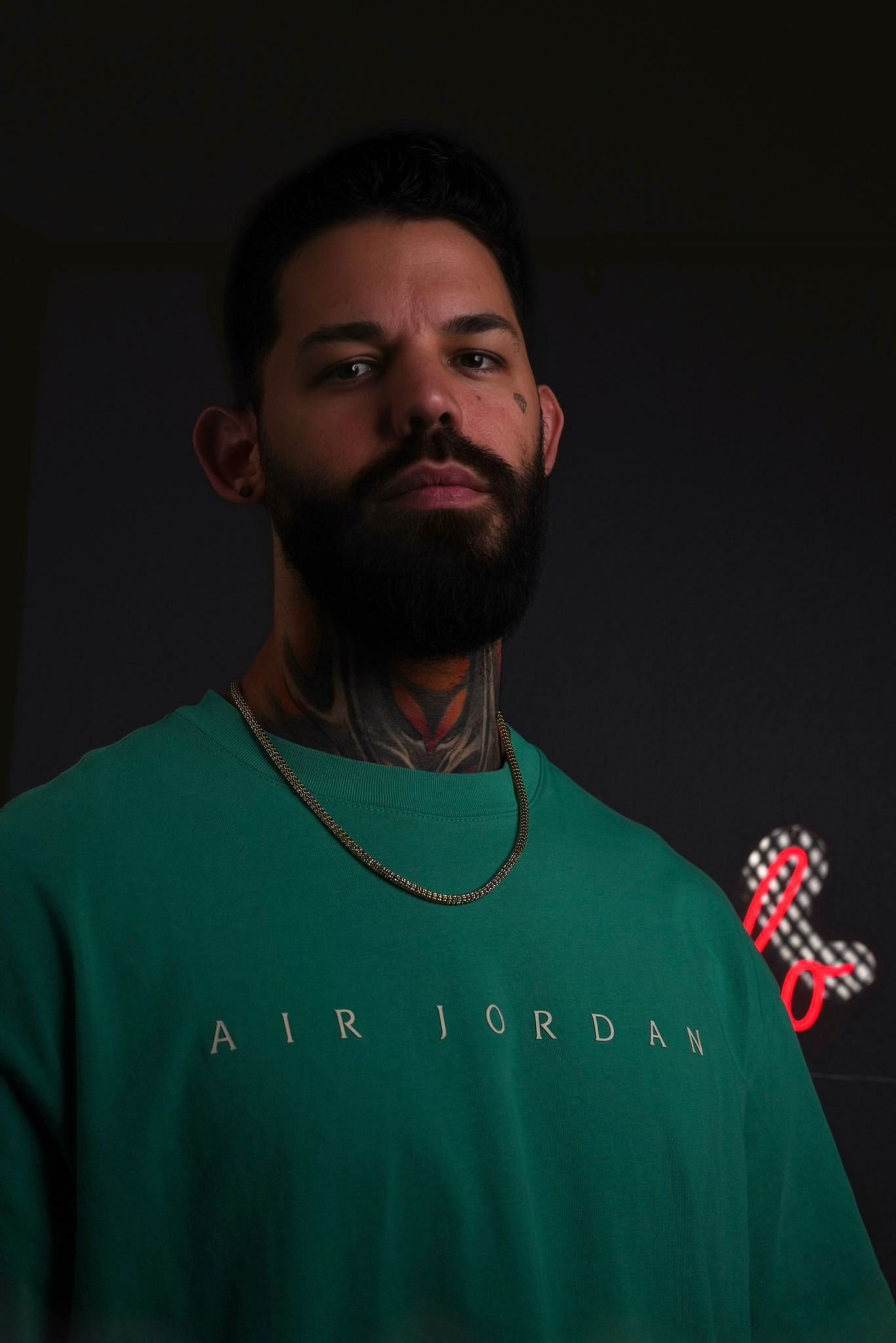 A man with a beard, tattoos, and a gold chain wears a green shirt with "AIR JORDAN" printed on it.